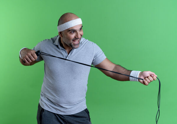 运动装戴着头巾 手拿跳绳 面带微笑的成熟运动型男子站在绿色的墙壁上微笑教练姿势
