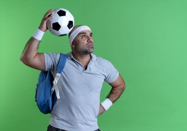 头带成熟的运动型男人戴着头巾 手里拿着足球 站在绿色的墙边茫然地看着一边足球站教练