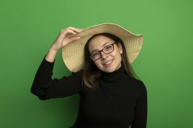 年轻年轻漂亮的女人戴着夏天的帽子 穿着黑色高领毛衣 戴着眼镜 快乐而积极地站在绿色的墙上 看着前面眼镜快乐女孩