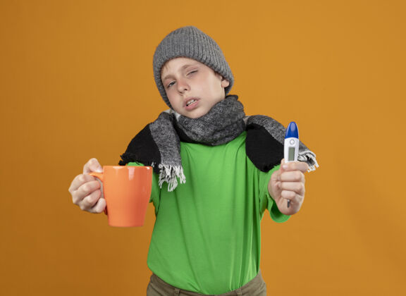 男孩生病的小男孩穿着绿色t恤 戴着暖和的围巾 戴着帽子 站在橙色的墙上 展示着一杯热茶和温度计 感觉不舒服 不舒服 不开心生病穿茶