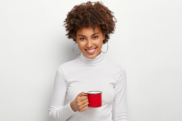 浓缩咖啡快乐的卷发女人的肖像 带着牙齿般的微笑 拿着红色的热饮杯 直视镜头休息满意享受
