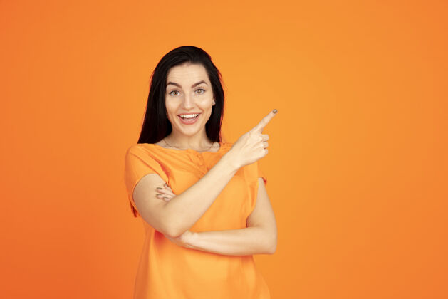 成功橙色工作室背景上的白种人年轻女子肖像穿着衬衫的漂亮黑发女性模特人类情感的概念 面部表情 销售 广告广告空间指向 展示 微笑人看起来衬衫
