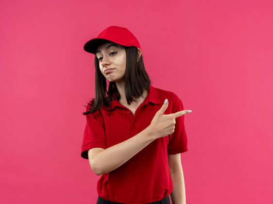 指向穿着红色马球衫 戴着帽子的年轻送货女孩看起来很不高兴 用食指指着站在粉红色墙上的一边年轻手指交货