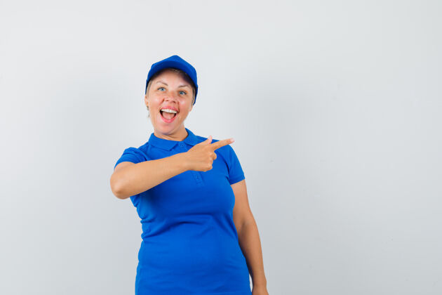 信心穿蓝色t恤的成熟女人指着旁边 看起来很高兴女性壁板帽子