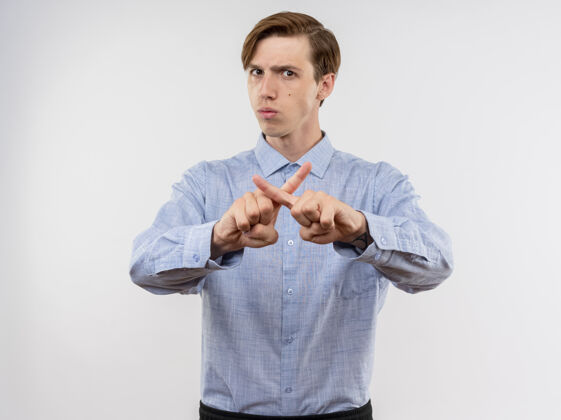 手指穿蓝衬衫的年轻人站在白墙上 用食指交叉做着防御的手势 严肃的脸目录男人年轻人