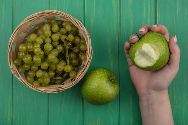 咬俯视图：在绿色的背景上 一个女人手里拿着绿色的苹果 一个篮子里有被咬过的绿色葡萄篮子观食物