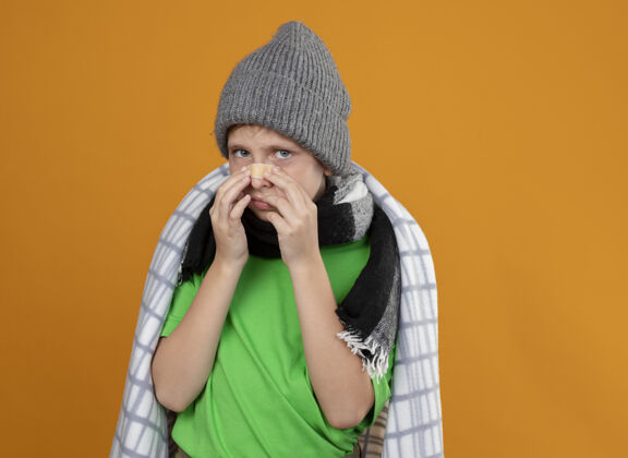 暖和生病的小男孩戴着暖和的帽子 围着围巾 裹着毯子 鼻子上贴着补丁 站在橘色的墙上不高兴又生病补丁围巾鼻子