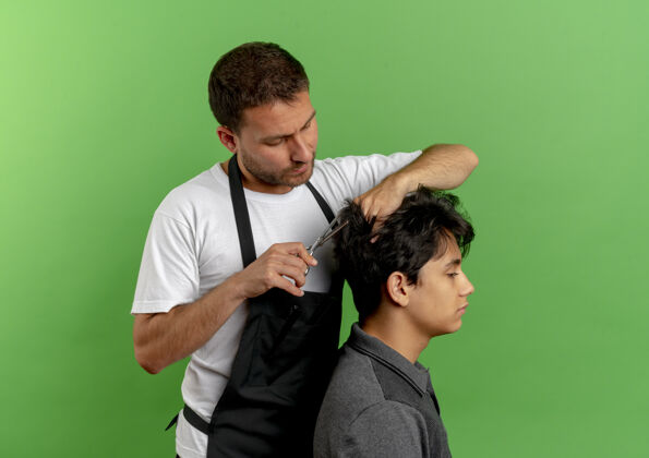 围裙穿着围裙的理发师正在用剪刀剪头发 满意的顾客站在绿色的墙上3头发人男人