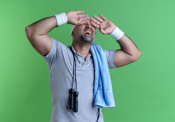 教练戴着头巾 脖子上缠着毛巾 手捂着眼睛 脸上带着恼怒的表情站在绿色的墙上的成熟的运动型男人脖子运动员烦恼