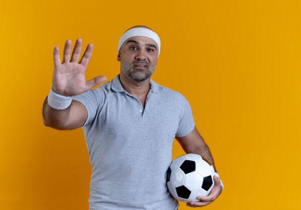 看戴着头巾的成熟运动型男子举着足球望着前方 手掌站在橙色的墙上运动教练运动