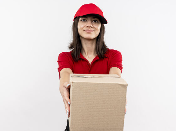 穿年轻的送货员穿着红色制服 戴着帽子 伸出盒子 友好地站在白色的墙上微笑着伸展制服包装