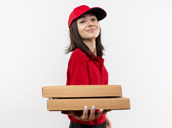 穿上年轻的送货员身穿红色制服 戴着帽子 手里拿着披萨盒 站在白色的墙上 脸上洋溢着幸福的笑容披萨站着拿着