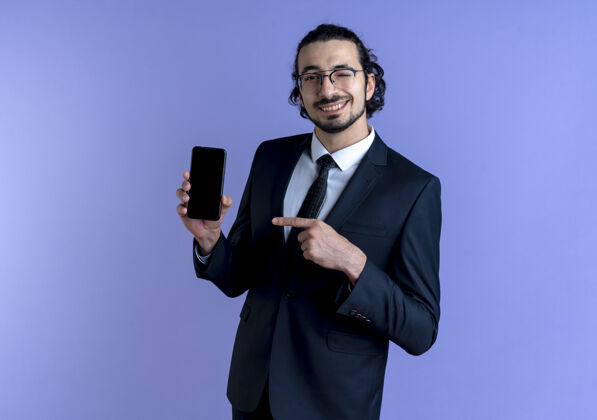眼镜一个穿着黑色西装戴着眼镜的商人站在蓝色的墙上 用手指着智能手机微笑着眨眼表演西装姿势