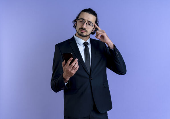 工作一位身穿黑色西装 戴着眼镜的商人看着智能手机屏幕 用手指着太阳穴 站在蓝色的墙上专注于一项任务商务站手指