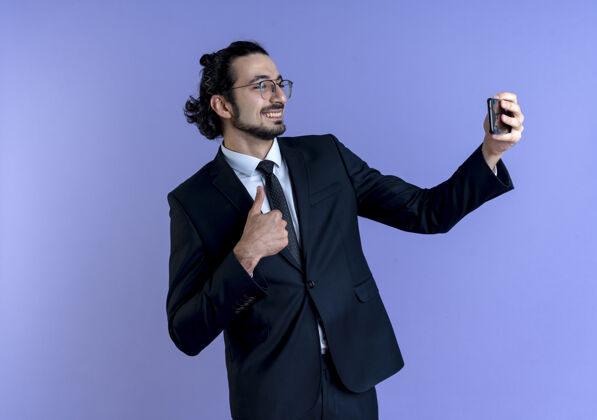 眼镜身穿黑色西装戴眼镜的商人用智能手机自拍微笑着竖起大拇指站在蓝色的墙上采取男人领带