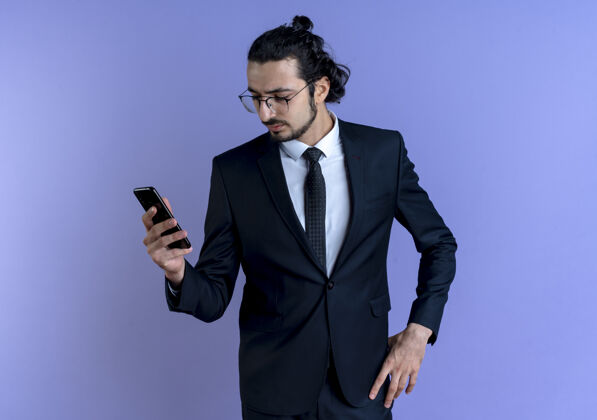 领带一个穿着黑西装戴着眼镜的商人站在蓝色的墙上 严肃地看着智能手机的屏幕姿势严肃商务