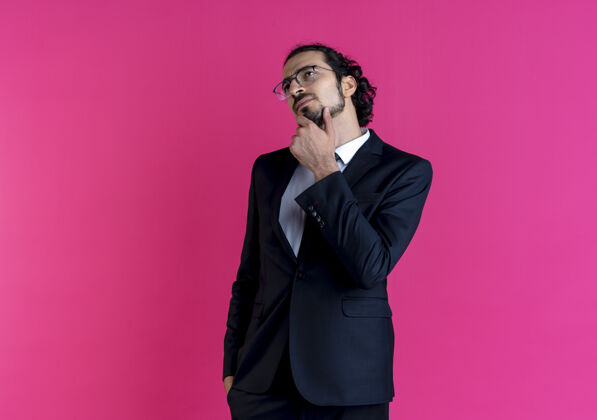 人一个穿着黑西装戴着眼镜的商人站在粉红色的墙上 手放在下巴上 困惑地看着一边优雅眼镜男