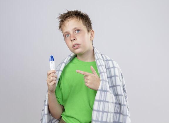不舒服生病的小男孩穿着绿色t恤 裹着毛毯 站在白色的墙上拿着温度计 用食指指着它 感觉不舒服小感觉男孩