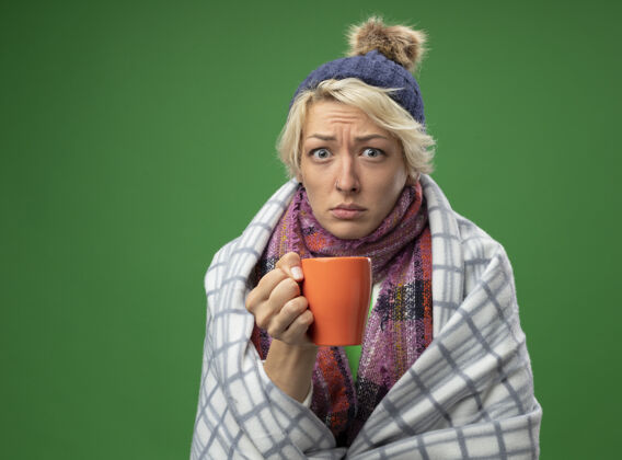 毯子生病的不健康的女人 短头发 戴着暖和的围巾和帽子 感觉不舒服 裹着毯子 端着一杯茶站在绿色的墙上包裹女人头发