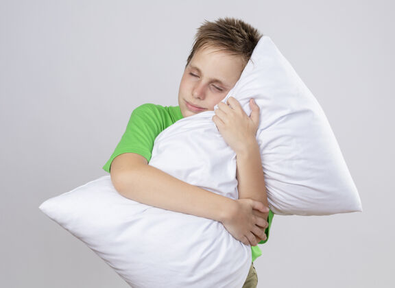 小生病的小男孩穿着绿色t恤感觉不舒服抱着枕头闭着眼睛站在白墙上男孩疾病枕头