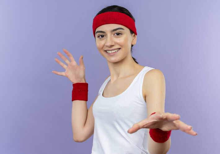 头带身穿运动服的年轻健身女士 头箍伸出手掌 站在紫色的墙上微笑着做防御手势人品牌年轻