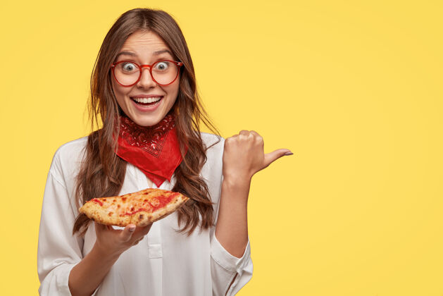 切片照片中表情激动的深棕色女士 带着牙齿般的微笑 戴着红色的头巾 拿着一片披萨 拇指放在一边 模特靠着黄色的墙壁为你做广告美味的菜肴右边吃快餐