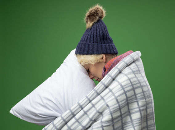 毯子生病的不健康的妇女 短头发 戴着暖和的围巾和帽子 感觉不舒服 裹在毯子里 抱着枕头 侧着身子 头靠在枕头上 背景是绿色的不适侧温暖
