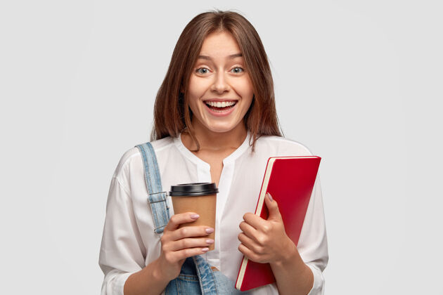 模特开朗的研究生穿着白衬衫 拿着外卖咖啡和笔记本 积极地微笑 隔着白墙女学员在室内做模特 满意吗工作服空闲时间候选人