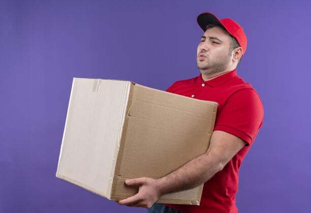 重量年轻的送货员身穿红色制服 头戴鸭舌帽 手里拿着一个大纸箱 站在紫色的墙上 看上去身体不适 身负重担盒子公民拿着