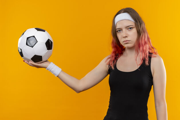 女人身穿运动服的年轻健身女士手拿足球 严肃的脸站在橙色的墙上健康人脸