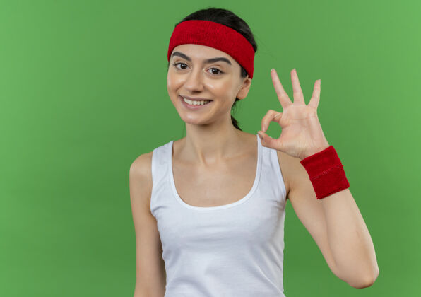 快乐身穿运动服的年轻健身女士 头上戴着一条快乐的头巾 面带乐观的笑容 站在绿色的墙上 高高兴兴地做着“ok”的手势健康市动