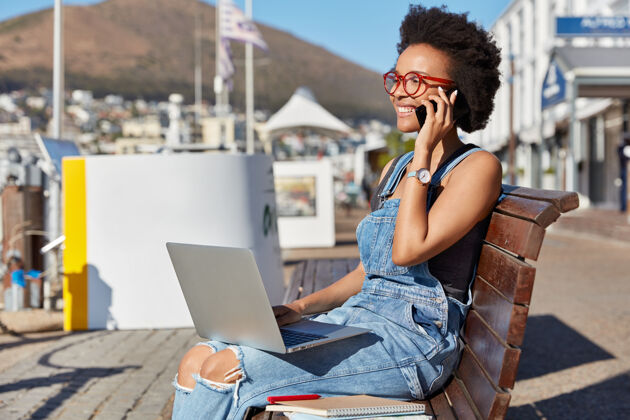 衣服照片中的美国黑人青少年微笑着通过手机给别人打电话 膝上放着笔记本电脑 坐在户外的长椅上 用小玩意儿上网学习 博客时尚 生活方式 科技工作服自由职业者当代