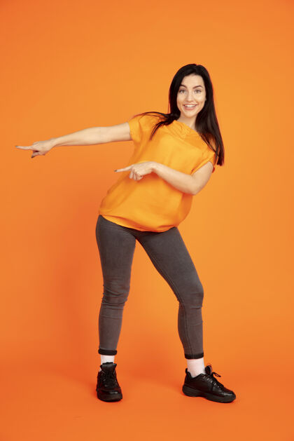 职业橙色工作室背景上的白种人年轻女子肖像穿着衬衫的漂亮深色女性模特人类情感的概念 面部表情 销售 广告广告空间指向 展示肖像员工表达
