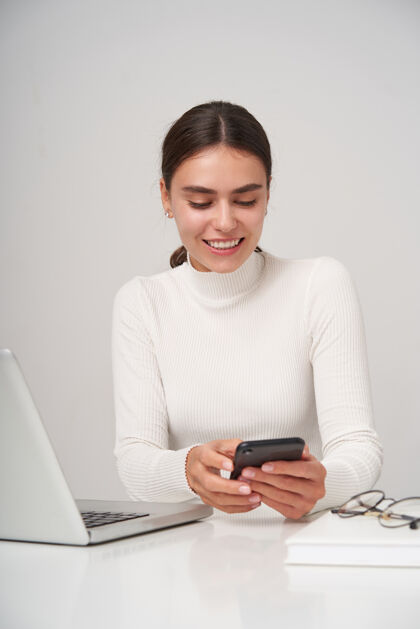 爱室内照片：快乐迷人的年轻黑发女性举着手机 积极地看着屏幕 读着朋友的留言女性笔记本电脑积极