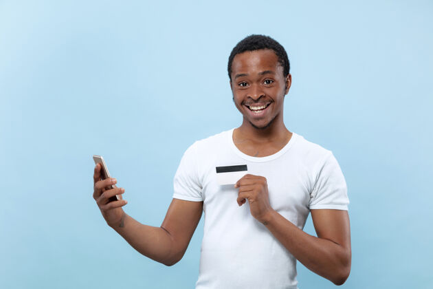 肖像身穿白衬衫 手持蓝色背景的卡片和智能手机的非洲裔美国青年半身像人类情感 面部表情 广告 销售 金融 在线支付概念行政人员在线胡须
