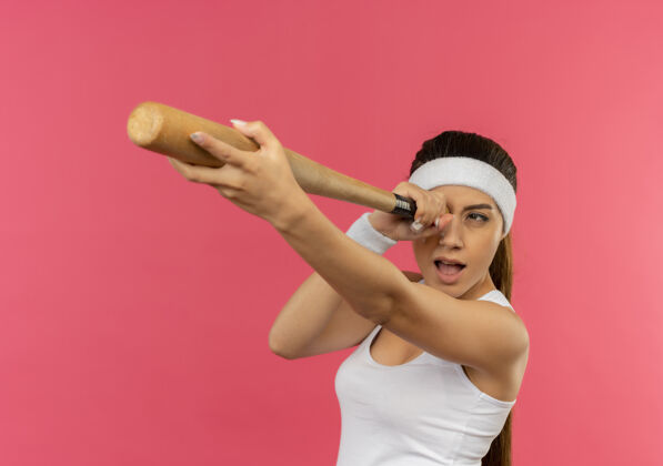 姿势身穿运动服的年轻健身女 头戴棒球棒 站在粉红色的墙上当望远镜年轻蝙蝠举行