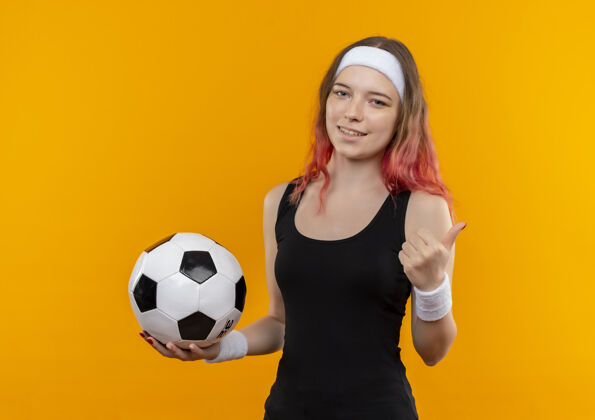 站着身着运动服的年轻健身女士站在橘色的墙上 手里拿着足球 脸上露出幸福的笑容 竖起大拇指拇指抱着足球