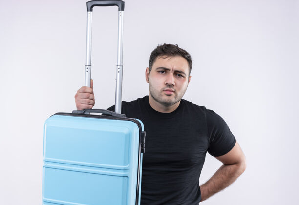 看着一个穿着黑色t恤的年轻旅行家站在白墙上 手里拿着一个皱着眉头的手提箱旅行旅行旅行者