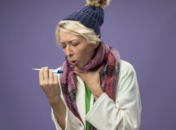 感觉生病的不健康的女人 短发 戴着暖和的围巾和帽子 感觉不舒服 拿着温度计 咳嗽 患流感 站在紫色的背景上不健康不适帽子