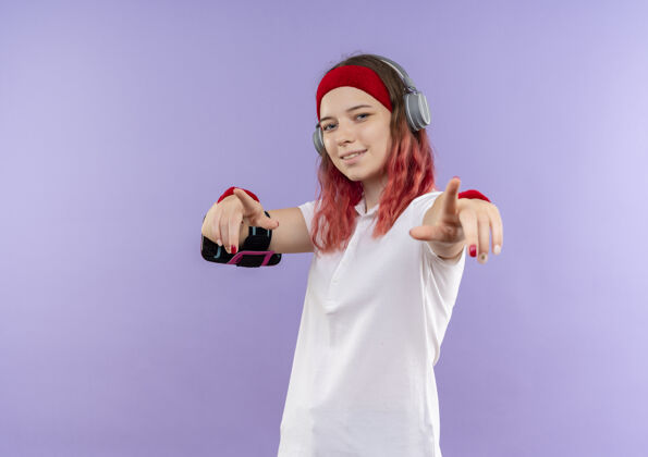 手指戴着头戴式耳机的年轻运动女性微笑着用手指指着摄像机 手持智能手机袖标站在紫色的墙上训练运动装运动微笑