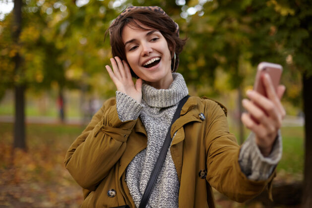 帽子活泼可爱的棕色头发的年轻女性 随意的发型 一边用手机举手 一边拍照 一边在泛黄的树上摆姿势 面带微笑姿势休闲年轻
