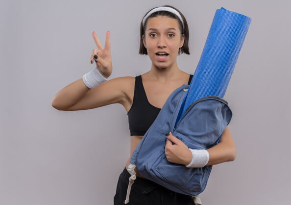 表演身穿运动服 背着背包 拿着瑜伽垫的年轻健身女士微笑着 在白色的墙上展示着胜利的标志微笑胜利垫子