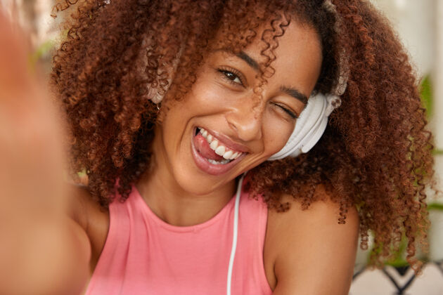 音乐积极卷曲的非裔美国女孩眨眼睛 露出舌头 心情好 用耳机听旋律 伸出手 用无法辨认的设备自拍肖像自拍非洲歌手