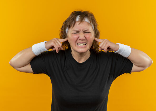 女性中年运动女性 身穿黑色t恤 戴着头巾 用手指捂着耳朵站在橙色的墙上 表情烦躁人女人站