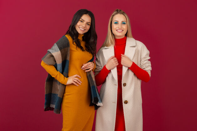 时尚两位笑容可掬的时尚女性穿着秋冬时装和外套 在红墙上孤零零地摆着姿势连衣裙衣服外套