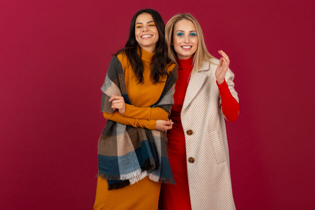 冷两位笑容可掬的时尚女性穿着秋冬时装和外套 在红墙上孤零零地摆着姿势女人时尚金发