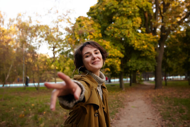 秋天积极的年轻迷人的黑发女士 随意的发型 微笑着愉快地伸出手 走过城市公园里的黄树女人姿势城市