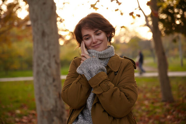 女士年轻漂亮迷人的棕色头发的女性 在穿过城市公园的时候 举手抚摸着脸颊 愉快地微笑着 穿着时髦的保暖服编织头发漂亮