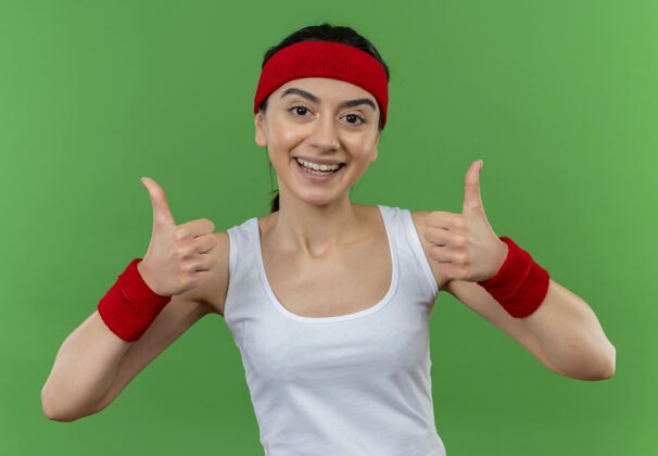 头带身穿运动服的年轻健身女士 头戴花环 快乐而积极地微笑着 站在绿色的墙上 愉快地竖起大拇指看运动健康