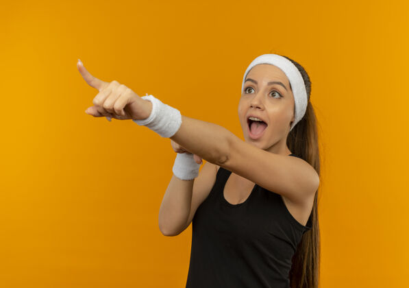 手指穿着运动服的年轻健身女 戴着头巾 手指着站在橘色墙上的某个令人惊讶的东西年轻人运动姿势